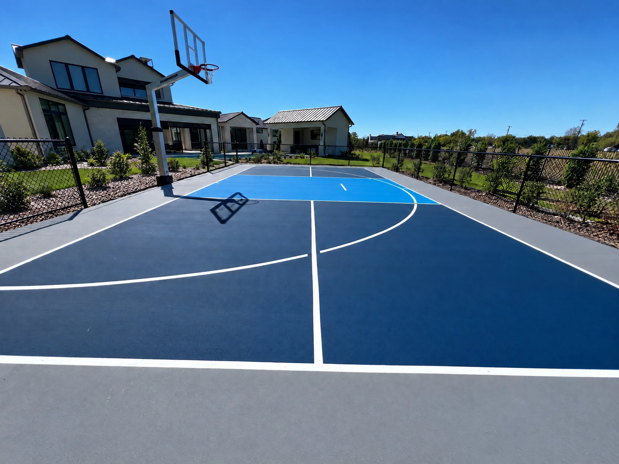 Blue and grey residential multi-sport court with white line markings, a basketball hoop, and perimeter fencing beside a modern home