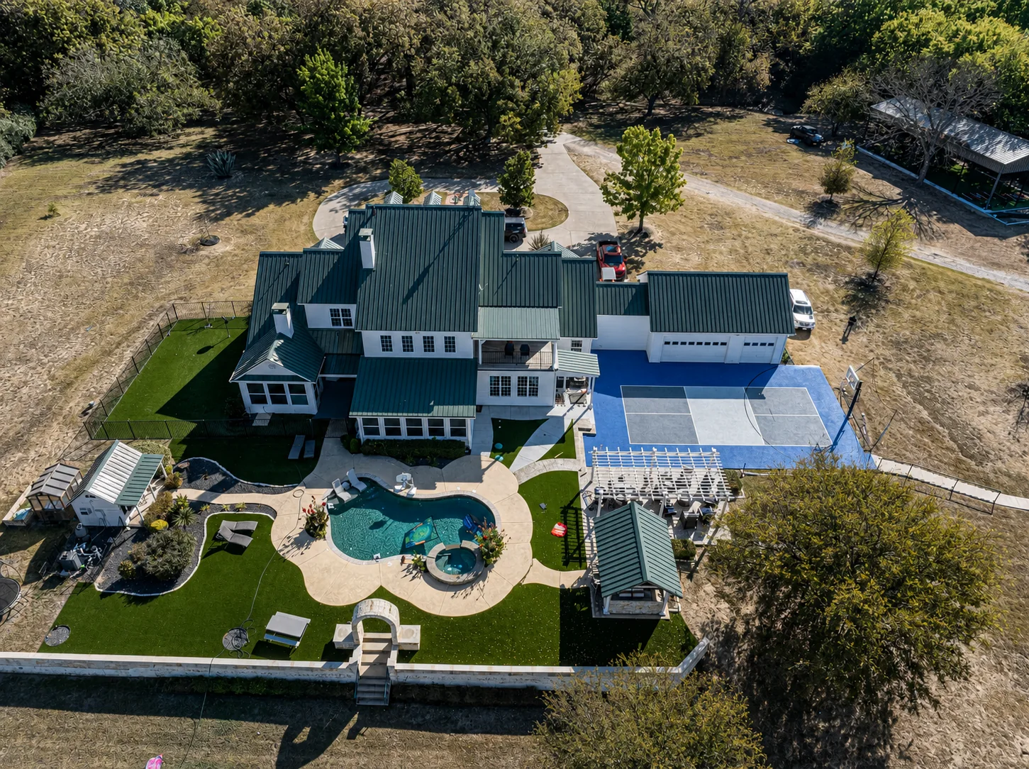 Wide aerial view of a green-roofed estate with a blue multi-sport court, pool, turf, and pergola