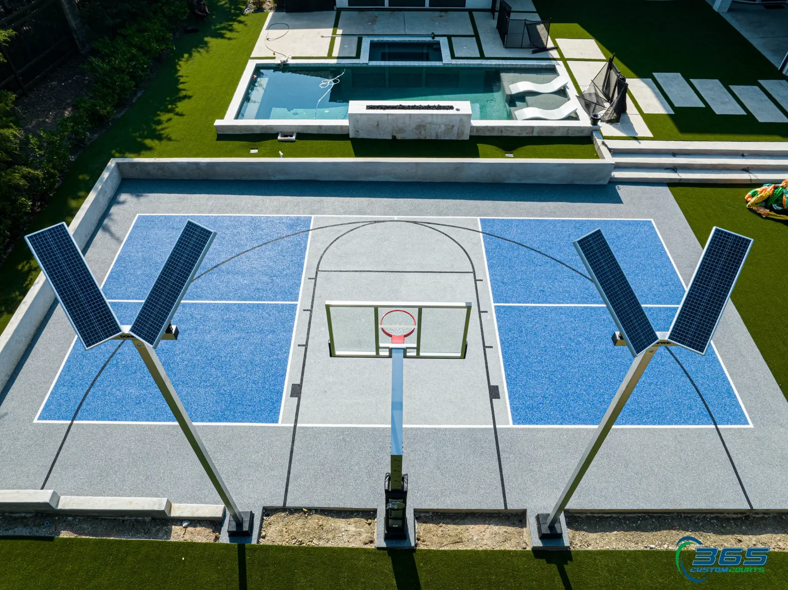Centered aerial view of a blue-and-grey multi-sport court with solar lights and a pool