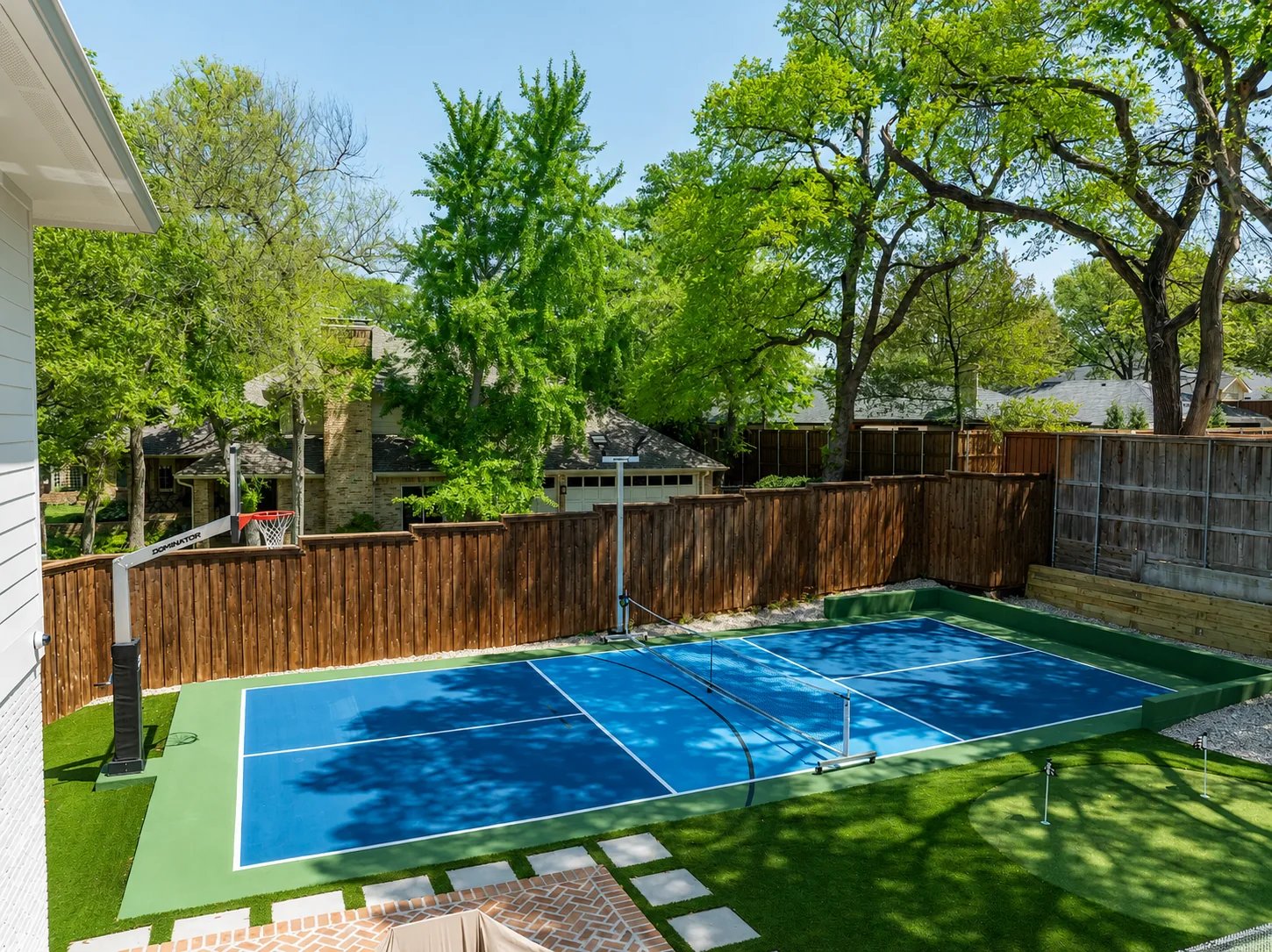 Blue backyard pickleball court framed by turf, fencing, and mature trees